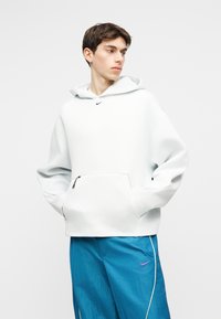 Ung mand iført en hvid hættetrøje og blå træningsbukser med lilla Nike-logoer, stående med hænderne i lommerne, ser mod højre.