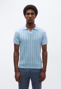 Ανοιχτό μπλε πλεκτό πόλο πουκάμισο με κάθετη ριγωτή υφή, κοντά μανίκια και γιακά με εγκοπή, συνδυασμένο με σκούρο παντελόνι.