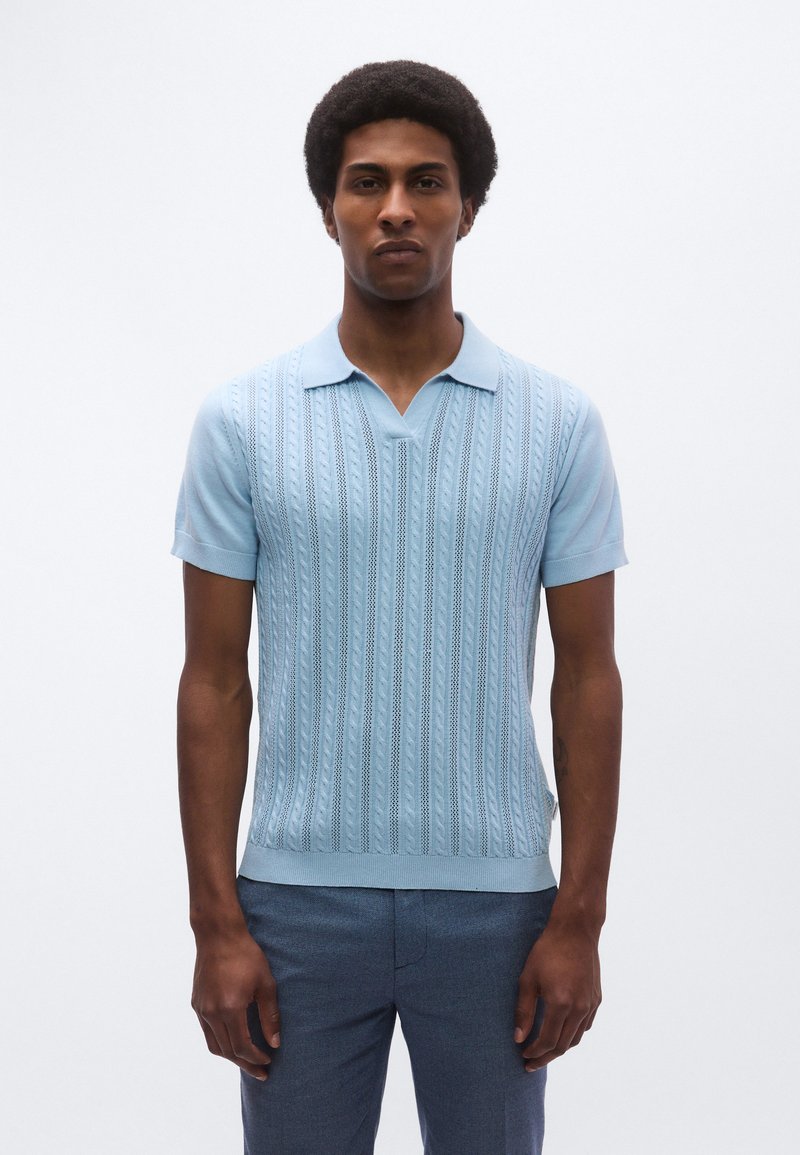 Ανοιχτό μπλε πλεκτό πόλο πουκάμισο με κάθετη ριγωτή υφή, κοντά μανίκια και γιακά με εγκοπή, συνδυασμένο με σκούρο παντελόνι.