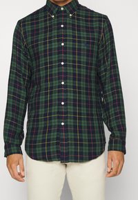Green plaid button-up shirt featuring a classic collar, white buttons, and contrasting red and yellow lines, made from a textured fabric.
