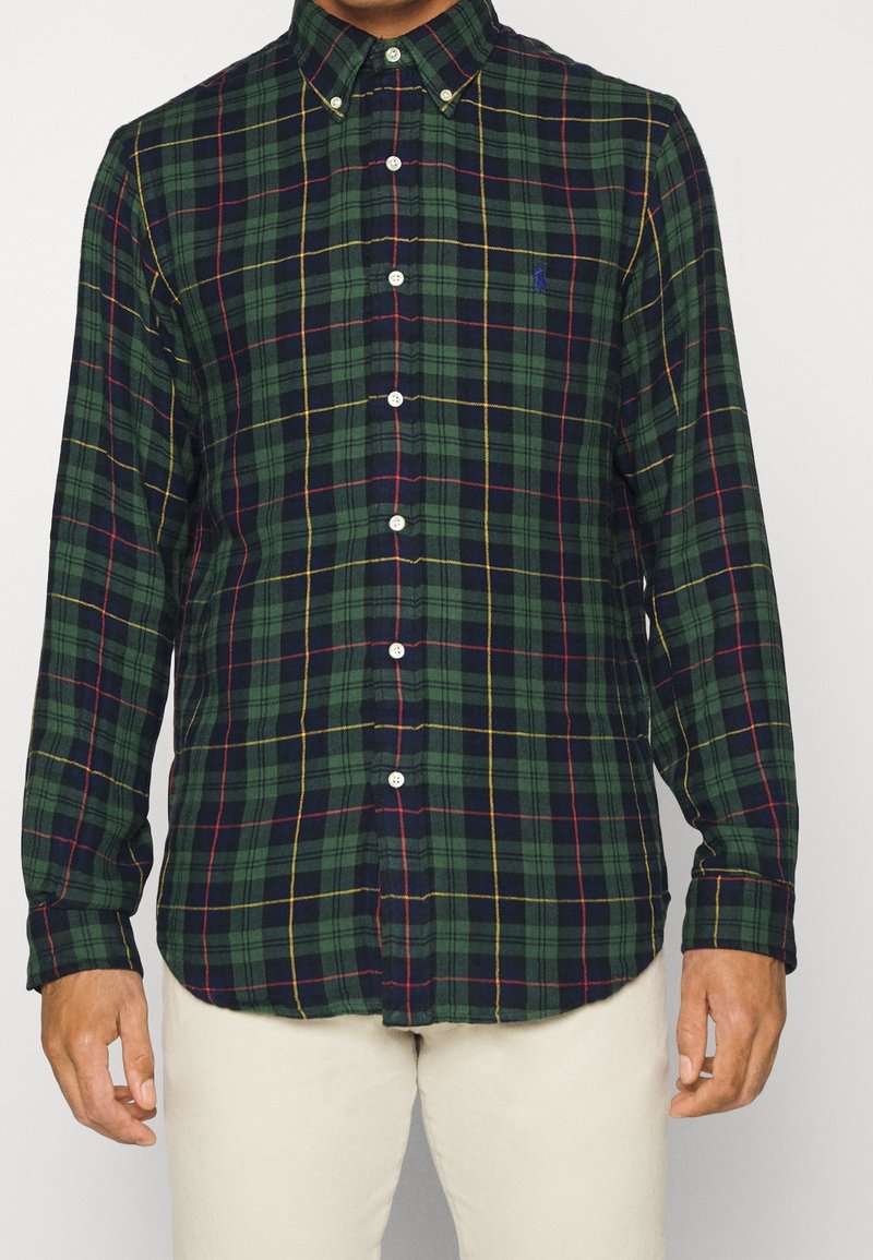 Green plaid button-up shirt featuring a classic collar, white buttons, and contrasting red and yellow lines, made from a textured fabric.