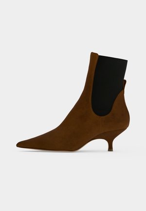 Stivaletto alla caviglia in camoscio marrone con punta appuntita, pannello elastico laterale nero e tacco kitten basso su uno sfondo chiaro uniforme.