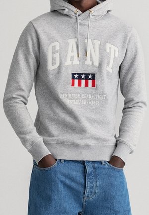 Mann trägt einen hellgrauen Kapuzenpullover mit "GANT" und einem rot-weißen-blauen Emblem, kombiniert mit blauen Jeans. Hände in den Taschen.