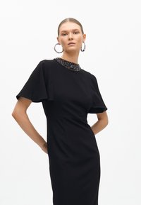 Svart klänning med korta utsvängda ärmar, dekorerad halsringning, figursydd silhuett, slät textur, som når till mitten av vaden.