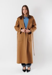 Cappotto in lana marrone con silhouette lunga, colletto ampio, due tasche frontali e una cintura. Indossato sopra un maglione blu a coste e jeans. Scarpe nere.