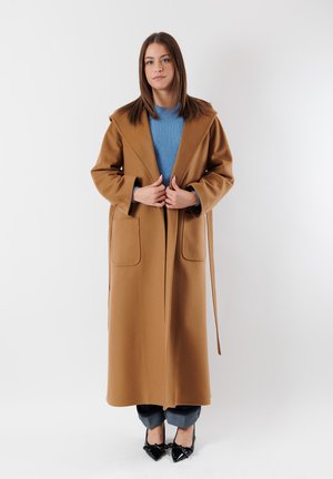 Cappotto in lana marrone con silhouette lunga, colletto ampio, due tasche frontali e una cintura. Indossato sopra un maglione blu a coste e jeans. Scarpe nere.