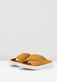Sandales à glisser en fausse suédine marron avec un design de lanières croisées, semelle en caoutchouc blanche et semelle intérieure orange. Apparence minimaliste et moderne.