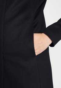 Černý kabát s texturovaným povrchem a boční kapsou. Obsahuje dlouhé rukávy a štíhlou siluetu se švy.