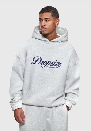 Jonge man die een lichtgrijze hoodie met het opschrift "Dropsize Studio Collection" en bijpassende joggingbroek draagt, kijkt naar links met zijn handen in de zakken.