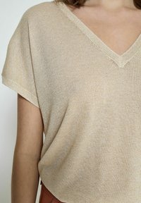 V-kaula, lyhythihainen beige paita. Pehmeä, teksturoitu kangas, jossa on hienovarainen kiilto ja rento leikkaus. Ompeleet kaula-aukon ympärillä.