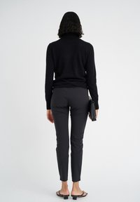 Schwarzer Rollkragenpullover, figurbetonte schwarze Hose und schwarze flache Sandalen. Die Person hält eine strukturierte schwarze Clutch und wird von hinten betrachtet.