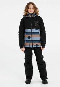 Jugendskijacke in Schwarz mit einem geometrischen Muster in Blau und Braun. Hat einen Reißverschluss vorne, einen hohen Kragen und ein Logodetail. Schwarze Hose und Stiefel.