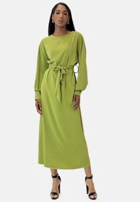 Robe verte à manches longues avec une taille nouée, une silhouette droite, une texture douce et un col rond, associée à des sandales à talons hauts noires.