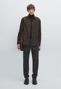 Chemise marron à boutons sur un col roulé marron, accompagnée de pantalons gris foncé et de chaussures en cuir marron foncé. Un sac à bandoulière en cuir marron est inclus.