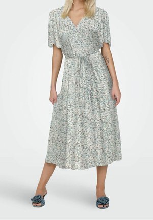 Femme portant une robe midi fleurie bleu clair à manches courtes et ceinture à la taille, associée à des sandales bleu clair à enfiler ornées de détails floraux en tissu.