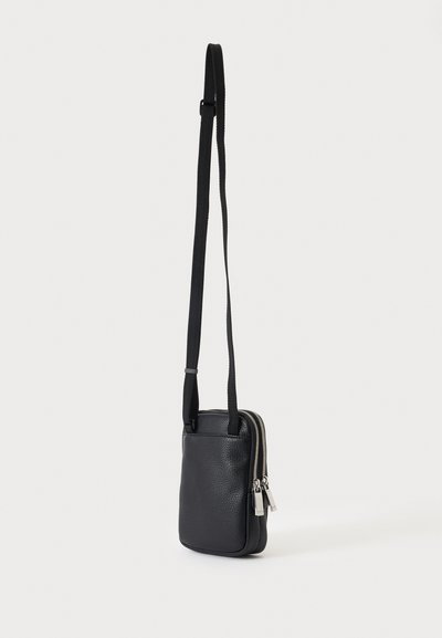 Sac bandoulière en cuir noir texturé avec deux fermetures éclair argentées et une longue bandoulière noire réglable sur un fond uni clair.