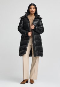 Czarna kurtka puchowa o błyszczącym wykończeniu, długości do kolan, z kapturem i zapinana na zamek. Nosi na sobie beżowy sweter dzianinowy i jasne spodnie.