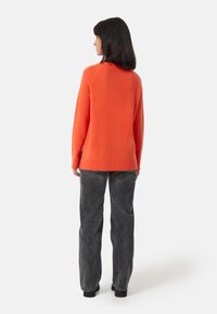 Maglione in maglia arancione brillante con scollo rotondo, maniche lunghe e orlo a costine, abbinato a jeans grigi a vita alta e scarpe nere.