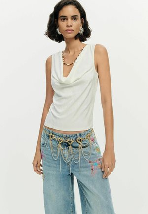 Žena s krátkymi tmavými vlasmi nosí biely bezrukávový top s kapucňovým výstrihom, modré džínsy s farebnou výšivkou a zlatý reťazový opasok s detailmi v tvare srdca.