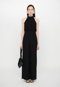 Svart halterneck jumpsuit med vide ben, med en fitted bodice og en glatt tekstur. Modellen holder en liten svart veske. Enkel design.