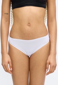 Femme portant un bas de bikini blanc et un crop top noir, montrant le ventre et un tatouage "DIVINE" sur son bras gauche.