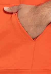 Felpa in cotone arancione con tasca e una mano all'interno, che mette in evidenza una texture liscia, un dettaglio della cucitura a forma di V e cuciture a contrasto.