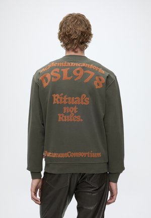 Osoba s kovrčavom kosom nosi zelenu majicu dugih rukava s narančastim tekstom "DSL 978", "Rituals not Rules" i drugim riječima na leđima.