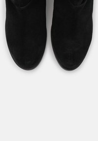 Une paire de bottes en daim noir avec des bouts arrondis photographiée d'en haut sur un fond clair.