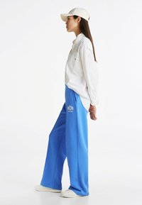 Niebieskie spodnie dresowe o luźnym kroju, z haftowanym logo. Noszone z białą koszulą na guziki i jasnym kapeluszem.