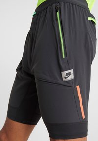 Närbild av svarta träningsshorts med neongrön dragkedjeficka, orange reflekterande rand och Nike-logotyp på låret.