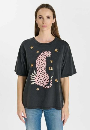 Frau trägt ein schwarzes T-Shirt mit einem pink gefleckten Leoparden und goldenen Sternen, kombiniert mit blauen Jeans, stehend vor einem schlichten Hintergrund.