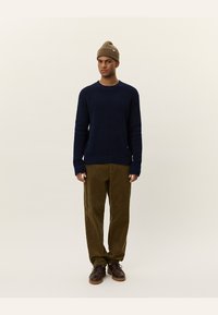 Navy knitted sweater with ribbed cuffs and hem, paired with olive corduroy pants. Model wears a brown beanie and brown lace-up shoes.