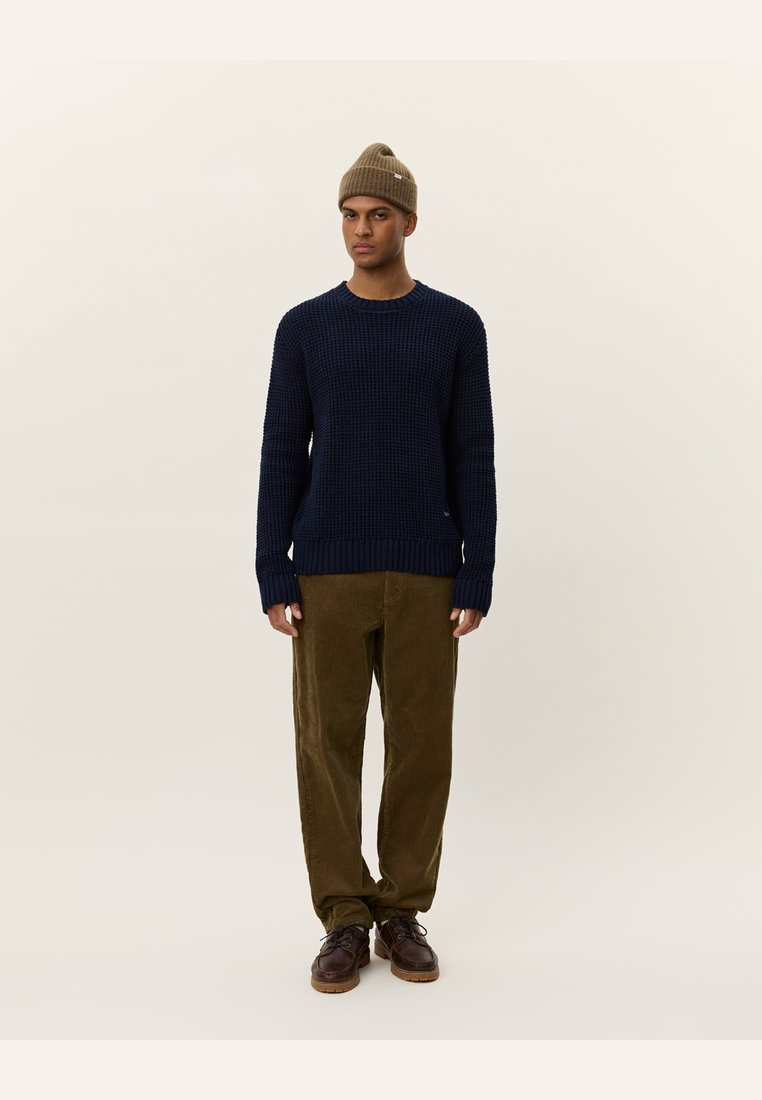 Navy knitted sweater with ribbed cuffs and hem, paired with olive corduroy pants. Model wears a brown beanie and brown lace-up shoes.