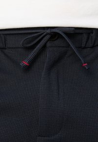 Morski hlače z teksturirano tkanino, ki imajo nastavljiv vrvico z rdečimi poudarki in zapenjalom na gumb. Opazne podrobne šive.