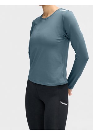 Kvinde iført en blå langærmet træningsshirt og sorte leggings med et lille hvidt logo på låret, der poserer med hænderne bag ryggen.