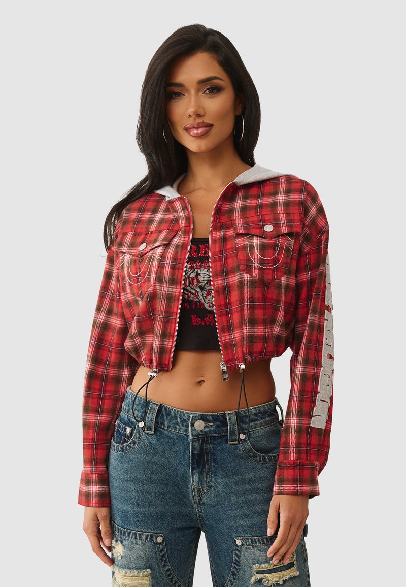 Vrouw in een rood geblokt cropped jack met een grijze capuchon, zwarte crop top en versleten blauwe spijkerbroek, staande tegen een effen achtergrond.