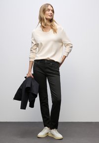 Beige svetr s výstřihem do V, černé slim-fit džíny, bílé tenisky, držící tmavou bundu, na světle šedém pozadí. Minimalistický design.