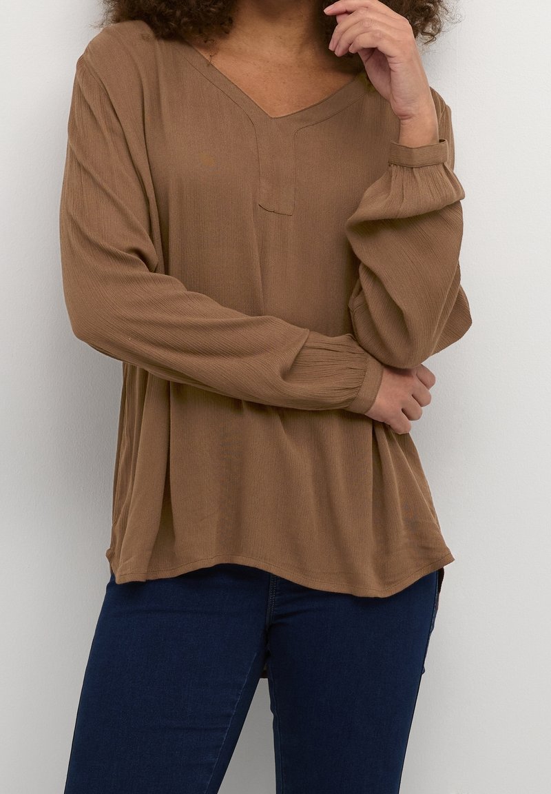 Bruine top met lange mouwen en V-hals, van textuurstof met gathered mouwen, gecombineerd met donkerblauwe jeans, weergegeven tegen een neutrale achtergrond.