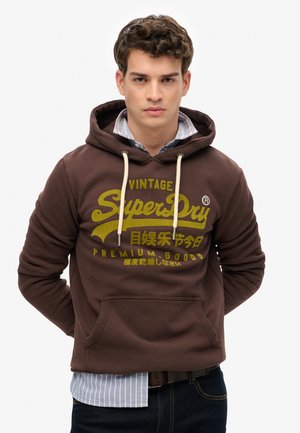 Unge mand iført brun Superdry hættetrøje med gul tekst og en blå stribet skjorte underneath, stående mod en ensfarvet hvid baggrund.