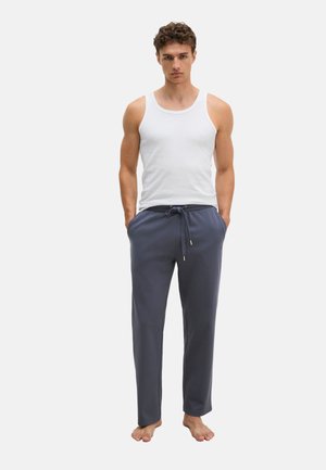Junger Mann steht barfuß, trägt ein weißes ärmelloses Shirt und graue Kordelzug-Hosen, die Hände in den Taschen, vor einem schlichten Hintergrund.