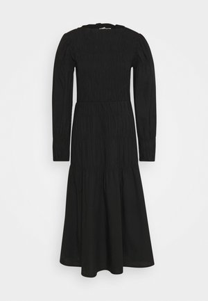 Lang sort kjole med puffede skuldre, rynket krop, tætsiddende talje, lange ærmer og udvidet skørt mod en ensfarvet hvid baggrund.