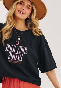 Černé oversized tričko s růžovým nápisem "HOLD YOUR HORSES." Má kulatý výstřih a krátké rukávy, doplněné o vrstvenou sukni.