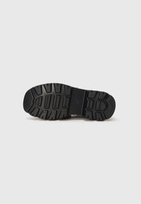 Black shoe sole with deep tread patterns, resting on a plain white surface, showing detailed grip design.