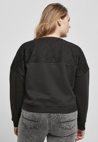 Felpa nera con schiena in pizzo testurizzato, parte anteriore uniforme e polsi a costine. Indossata con jeans grigio scuro. Design semplice e moderno.