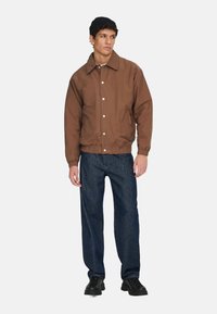 Braune Bomberjacke mit Kragen, elastischem Taillenbund und Druckknopfverschluss. Kombiniert mit dunkler Denimhose und schwarzen Schuhen.