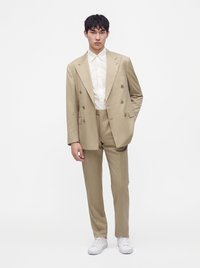 Giovane uomo in piedi che indossa un abito doppiopetto beige, camicia bianca con bottoni e sneakers bianche su uno sfondo bianco semplice.