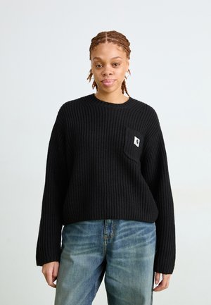 Schwarzer Rippstrickpullover mit rundem Ausschnitt, überschnittenen Schultern und einer Fronttasche mit einem Logo-Patch. Getragen mit hellblauen Jeans.
