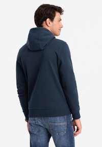 Felpa blu navy con cordino, realizzata in tessuto morbido, con polsini e orlo a coste, indossata con jeans azzurri chiari.