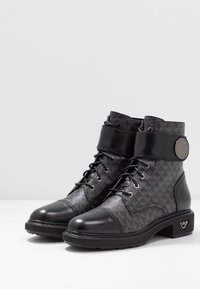 Černé kotníkové boty s texturovaným svrškem a logem, šněrování a lesklou lakovanou špičkou. Masivní podpatek s detaily loga.