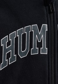 Svart sweatshirt med "HUM" i fet grå text, dragkedjedetalj på sidan och en strukturerad tygyta.